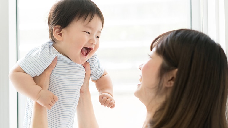 A smiling mother holds up her laughing baby in a bright room near a window.