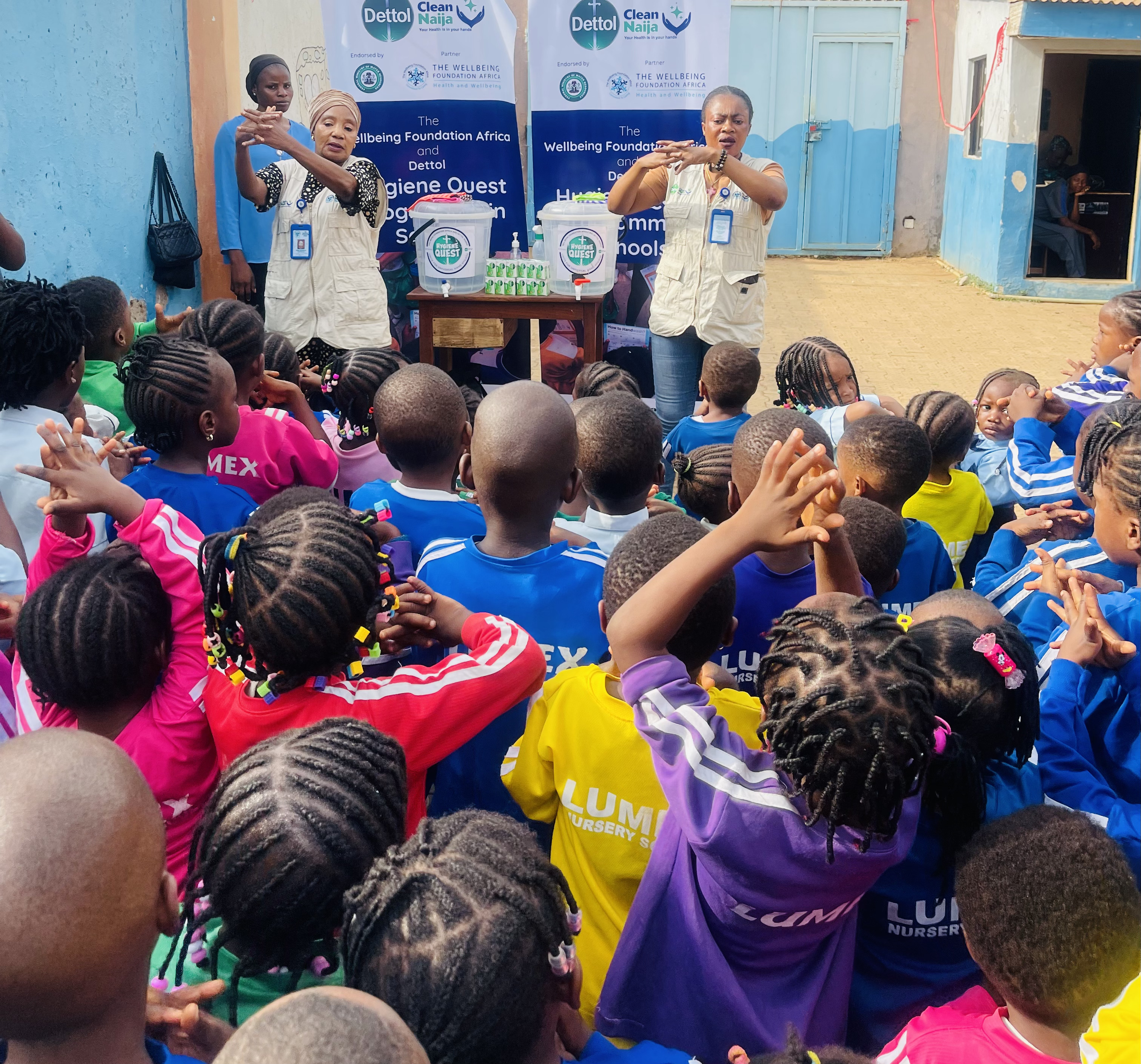 A group of children in colorful clothing are participating in a handwashing demonstration led by two instructors in front of banners promoting hygiene