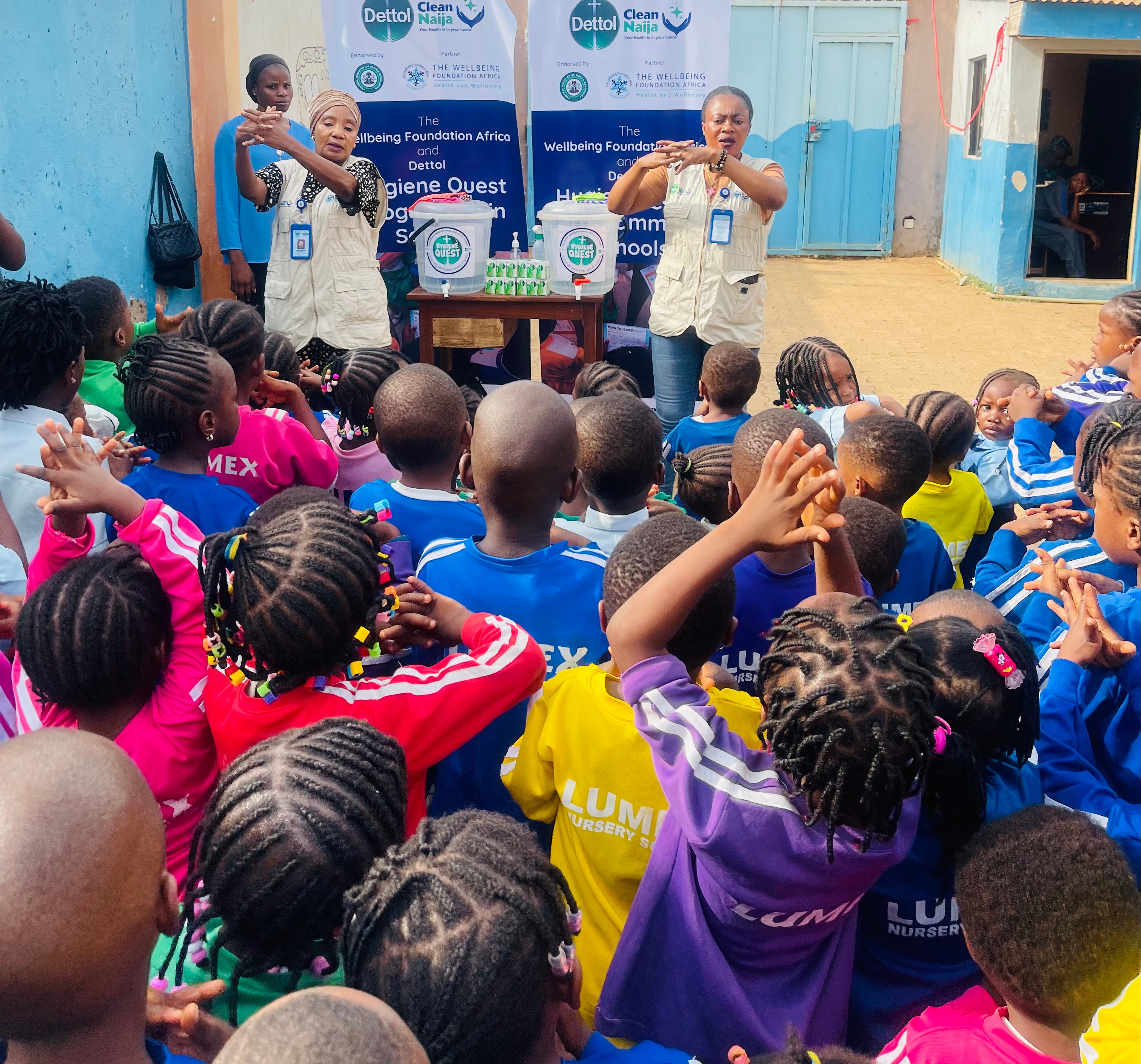 A group of children in colorful clothing are participating in a handwashing demonstration led by two instructors in front of banners promoting hygiene