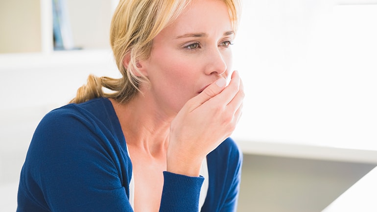 A blonde woman in a blue cardigan covers her mouth with her hand while checking her breath.