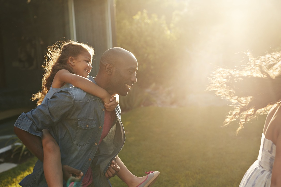 Man giving a piggyback ride to a young girl outdoors in a sunlit garden or backyard.