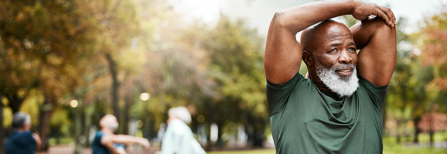 Older man stretching outdoors in a park with people exercising in the background