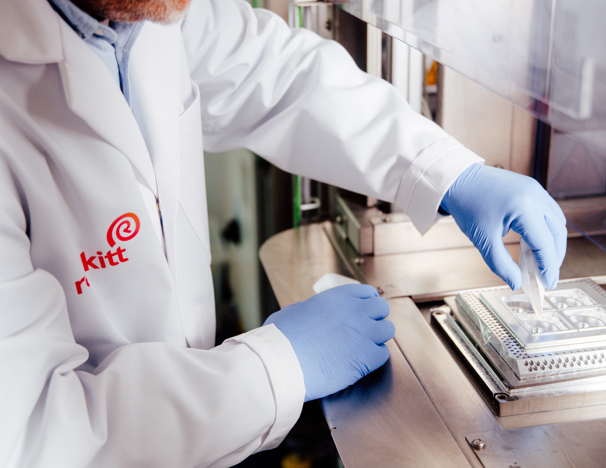 A person wearing a lab coat and blue gloves is handling a small container near laboratory equipment