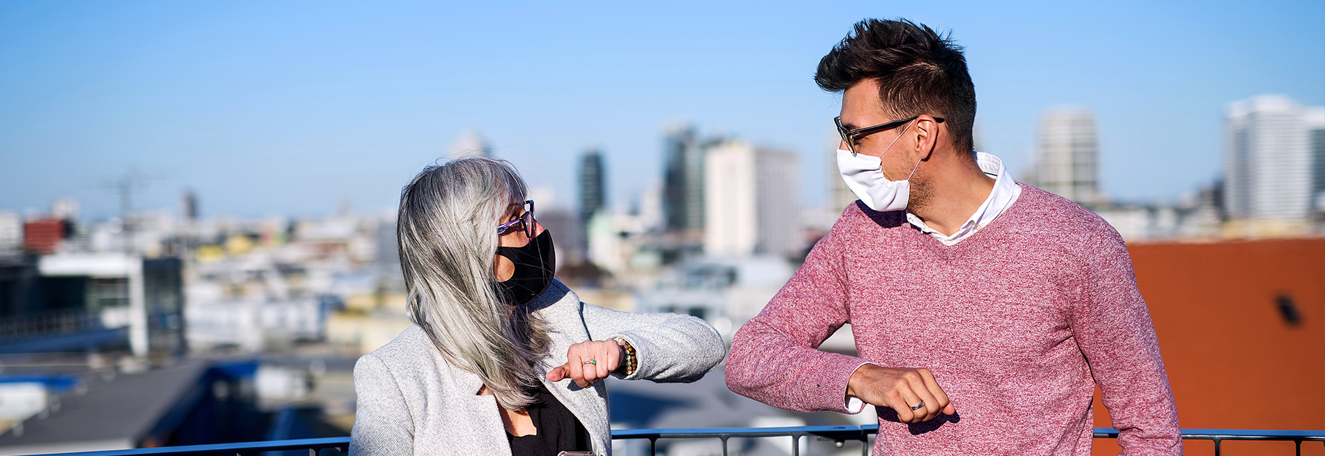 Two people wearing masks greet each other with elbow bumps on a rooftop with a city skyline background.