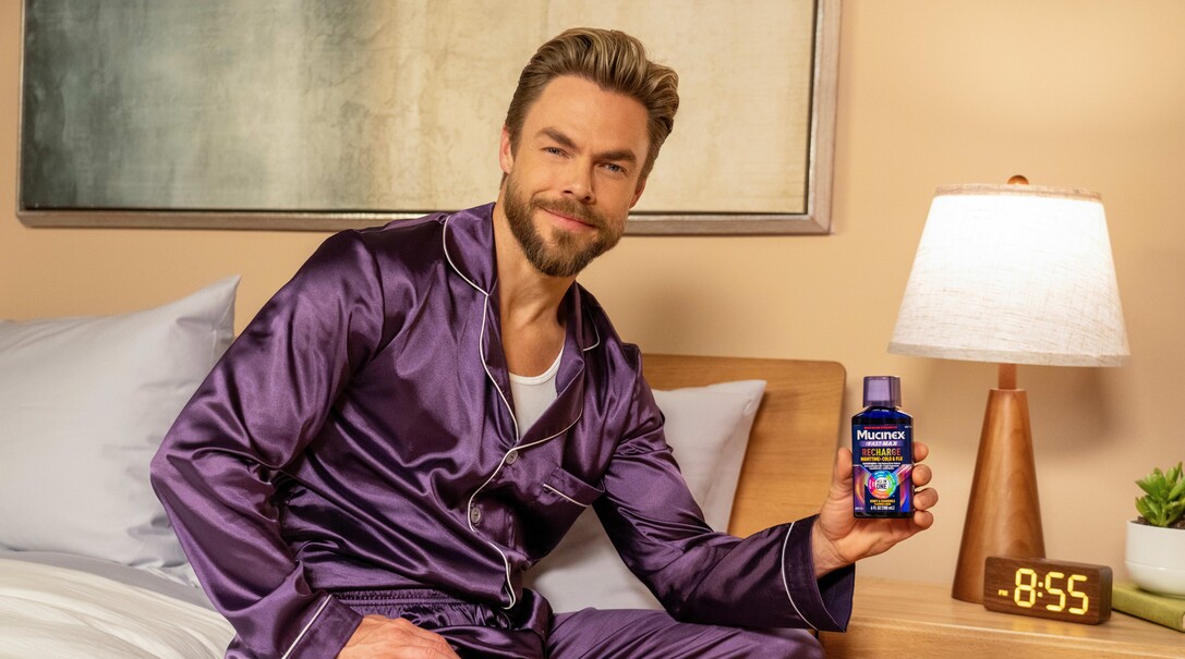 Derek Hough sitting on a bed in purple satin pajamas, holding a bottle of Mucinex Fast-Max Recharge next to a bedside lamp and clock.