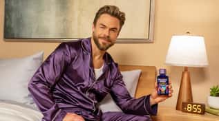 Derek Hough sitting on a bed in purple satin pajamas, holding a bottle of Mucinex Fast-Max Recharge next to a bedside lamp and clock.