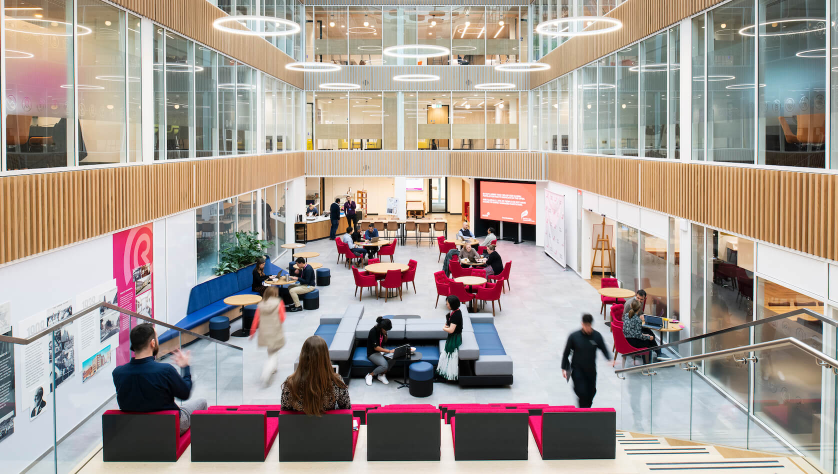 Open-plan office atrium with people collaborating, working, and relaxing in shared spaces.