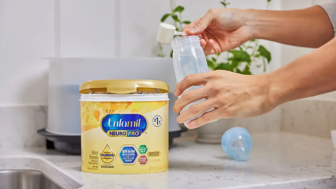 Hands preparing a baby bottle with Enfamil NeuroPro formula on a kitchen counter.