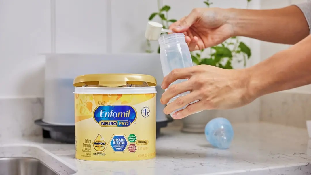 Hands preparing a baby bottle with Enfamil NeuroPro formula on a kitchen counter.