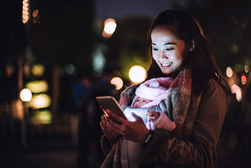 Woman holding a baby while looking at a phone on a city street at night