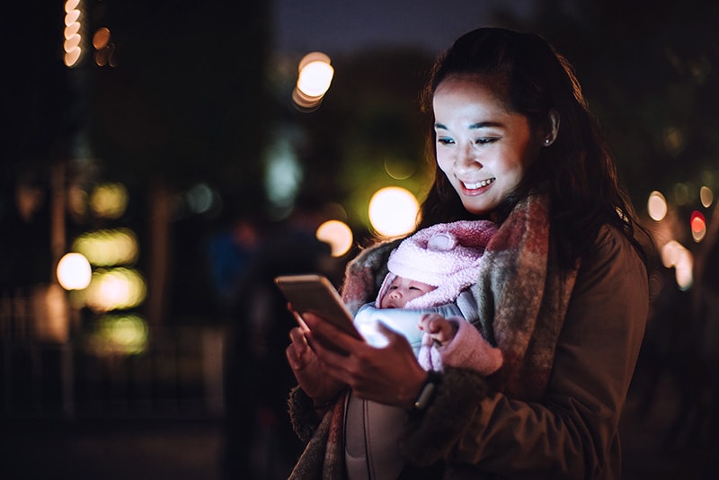 Woman holding a baby while looking at a phone on a city street at night
