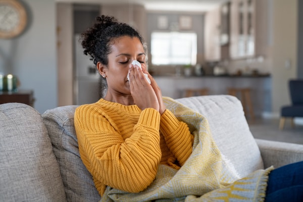 A woman sitting on a sofa, using a tissue to relieve sneezing in a cozy home setting