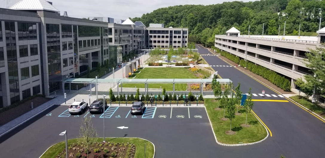 A large modern office complex with landscaped gardens, multiple parking spaces including handicapped spots, and a multi-level parking structure surrounded by greenery.