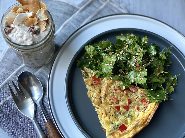 Slice of vegetable frittata topped with fresh herbs, served alongside an overnight oats parfait on a table setting