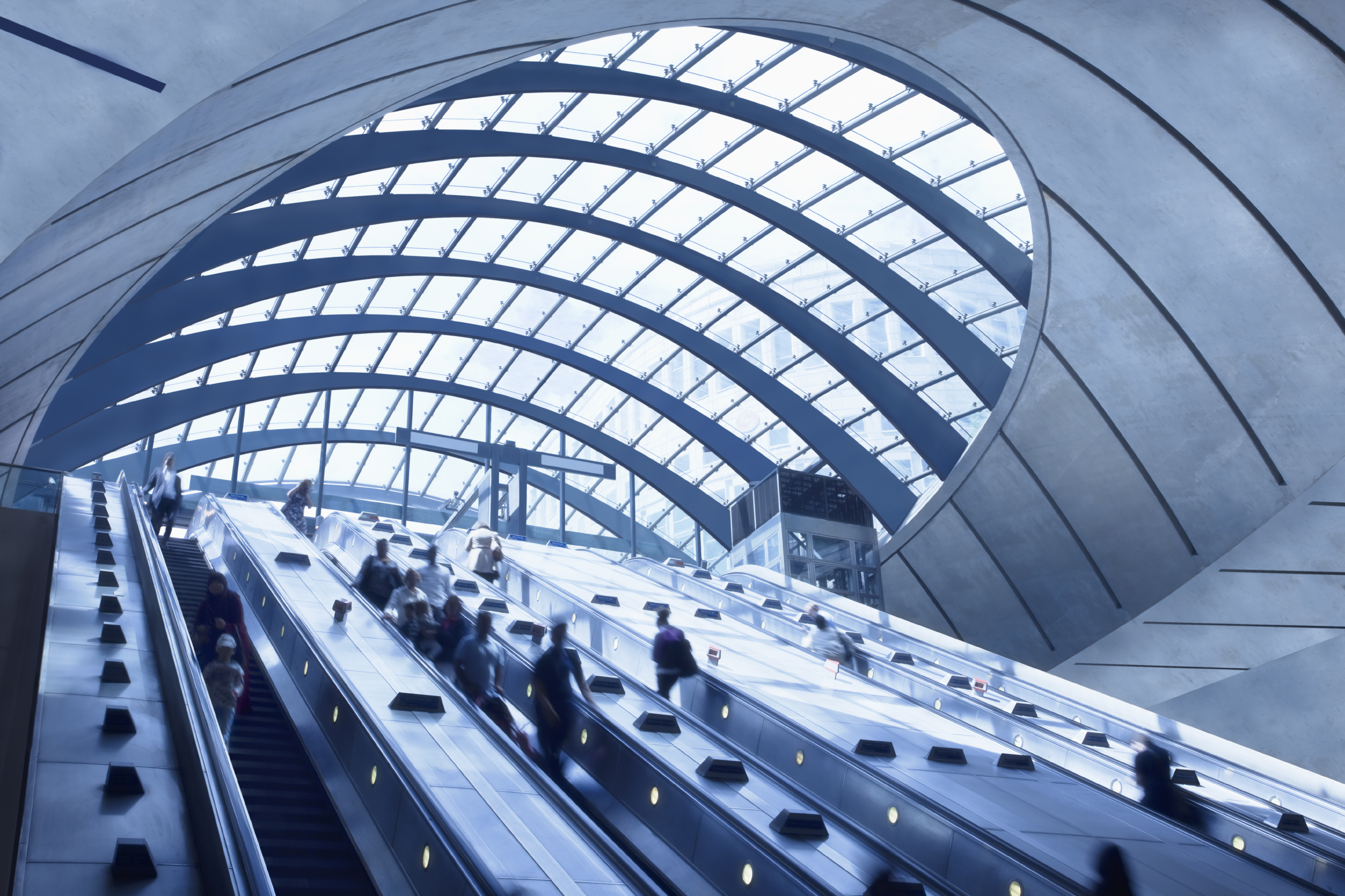People riding escalators inside a modern transit station with a curved glass ceiling