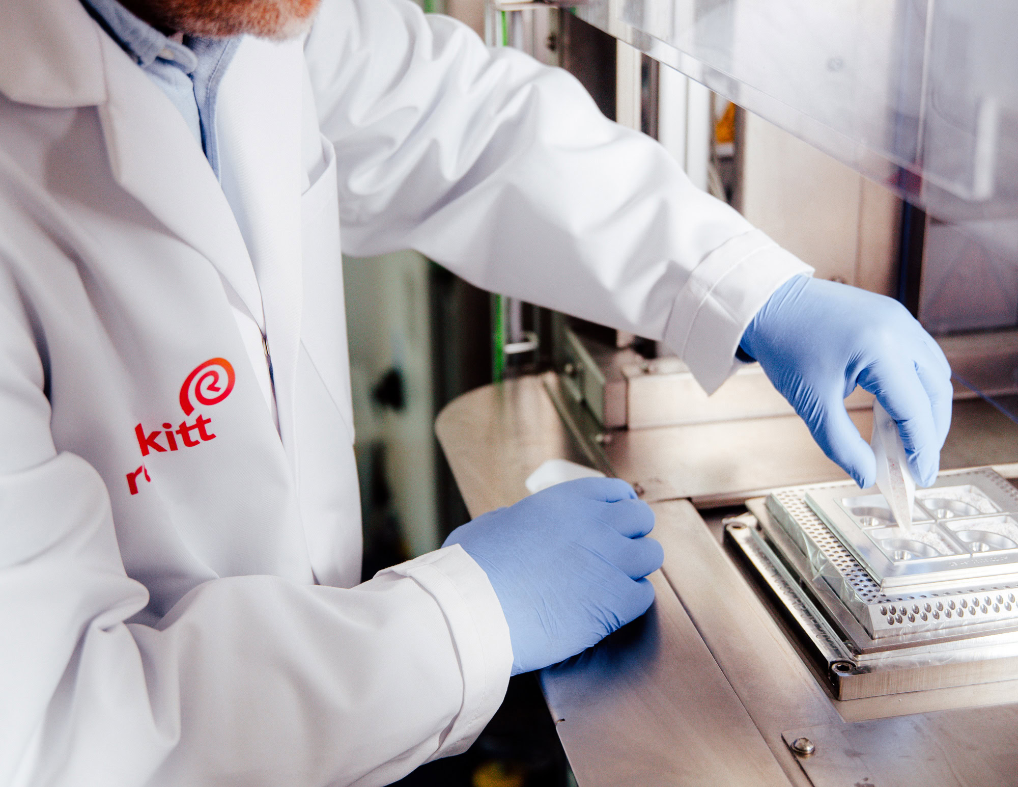 Gloved lab technician in a white coat placing samples with tweezers into a metal tray.