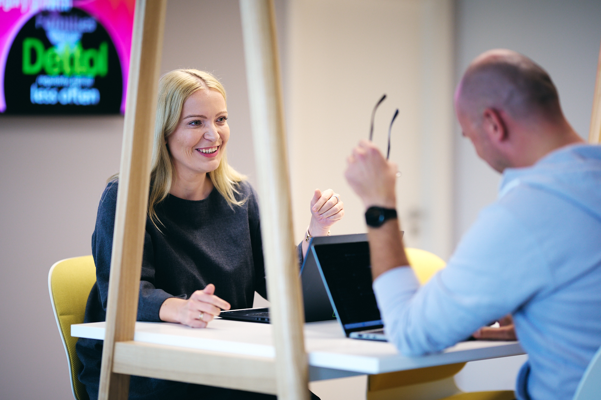 Two colleagues smiling and talking at a desk with laptops.
