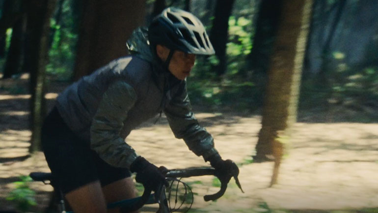 A cyclist wearing a helmet and a jacket rides a bicycle along a wooded trail.