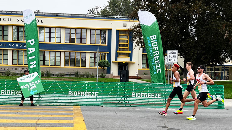 Three runners race along a street past green Biofreeze promotional banners and a high school building.