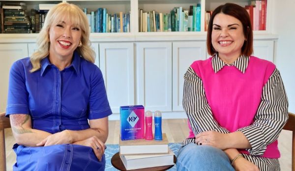 Two smiling women sit together in front of a bookshelf, with a package of K-Y Yours+Mine lubricants displayed on a small table between them.