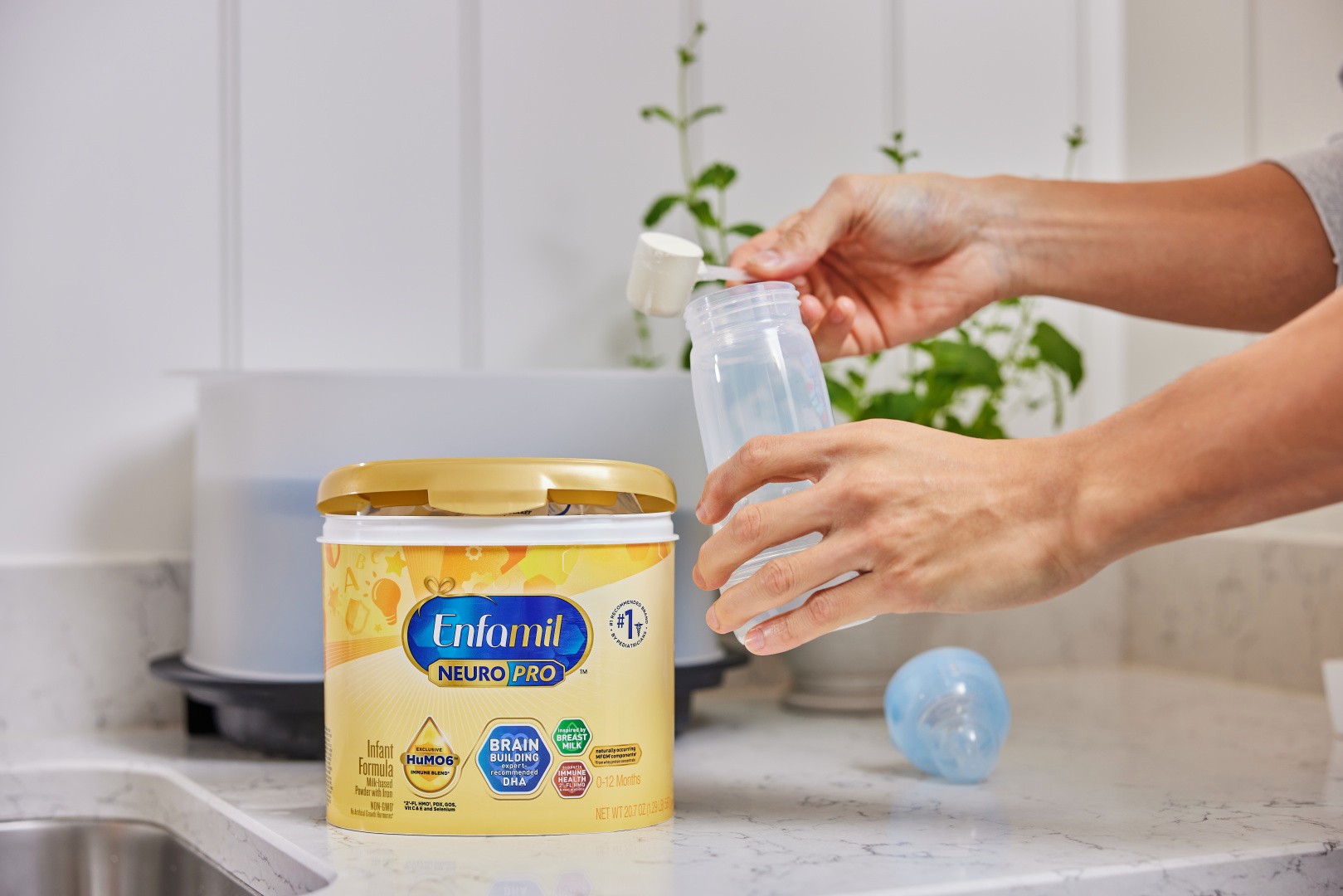A person scooping Enfamil NeuroPro infant formula powder into a baby bottle on a kitchen countertop.