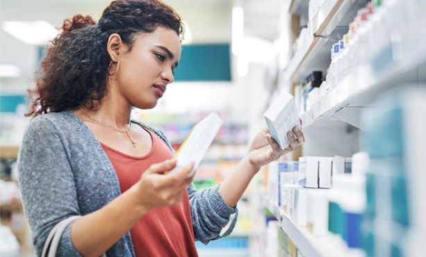 Woman comparing two product boxes while shopping in a store aisle with shelves full of various items.