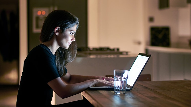 A person working on a laptop at a wooden table in a dimly lit kitchen with a glass of water nearby.