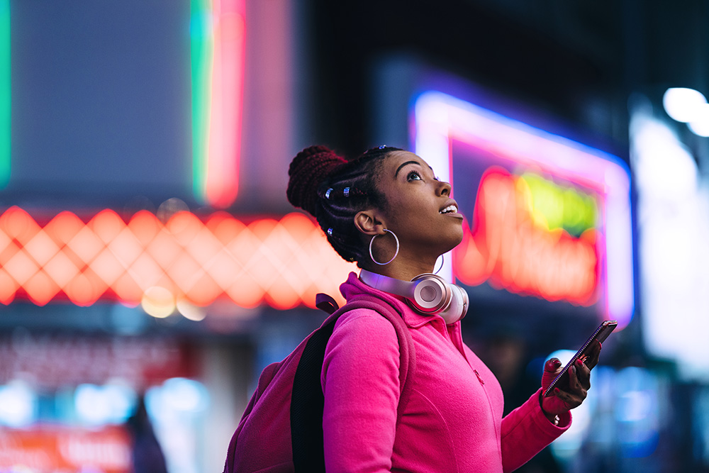 Woman with headphones and a phone standing in front of colorful neon city lights at night