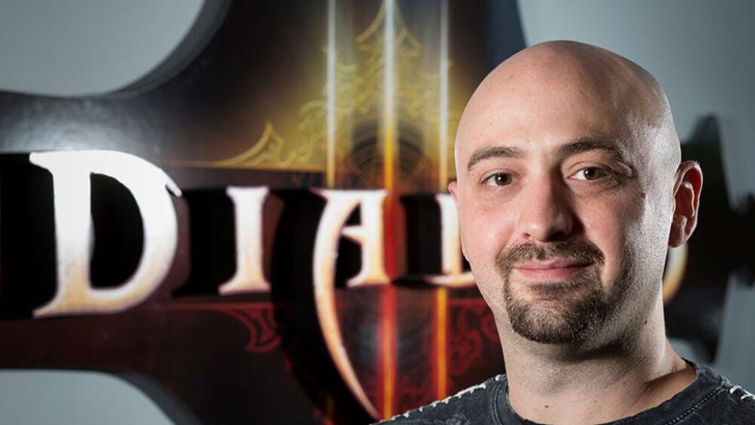 A photograph of Travis Day. He is bald, bearded, and wearing a black T-shirt while posing in front of a Diablo III poster.