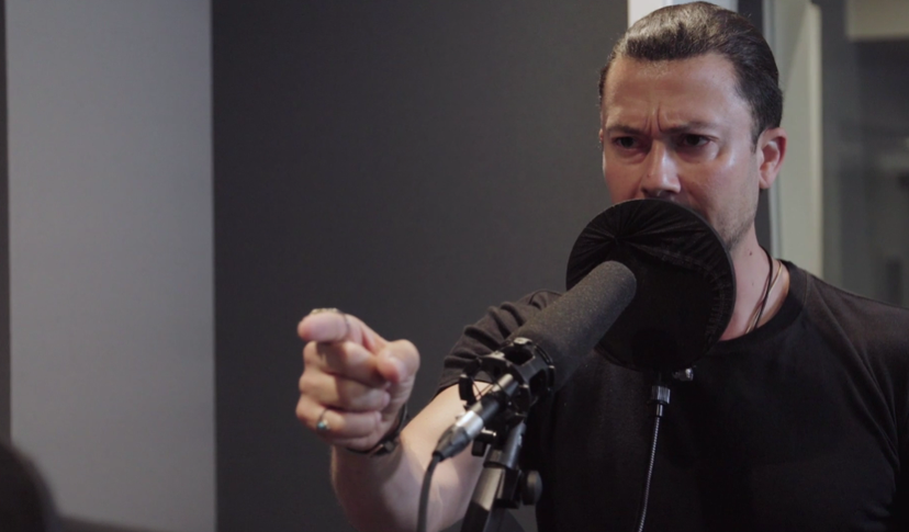 A man records voiceover work at a standing microphone. His expression is stern and his hand points offscreen.