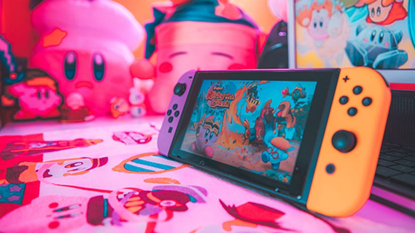 A photograph of a Nintendo Switch displaying the character of Kirby, surrounded by Kirby merch.