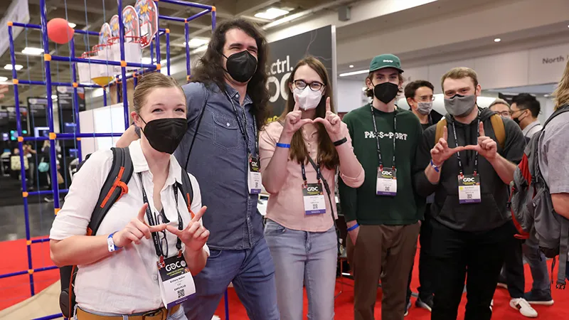 A group of GDC attendees wearing masks smile and make L symbols for the camera. 
