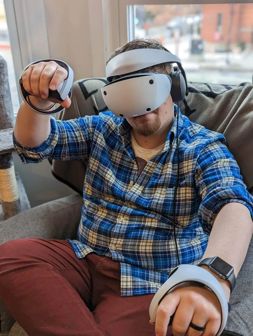 Bryant sits on a chair while using the PSVR2. He is wearing a blue striped shirt and red pants.