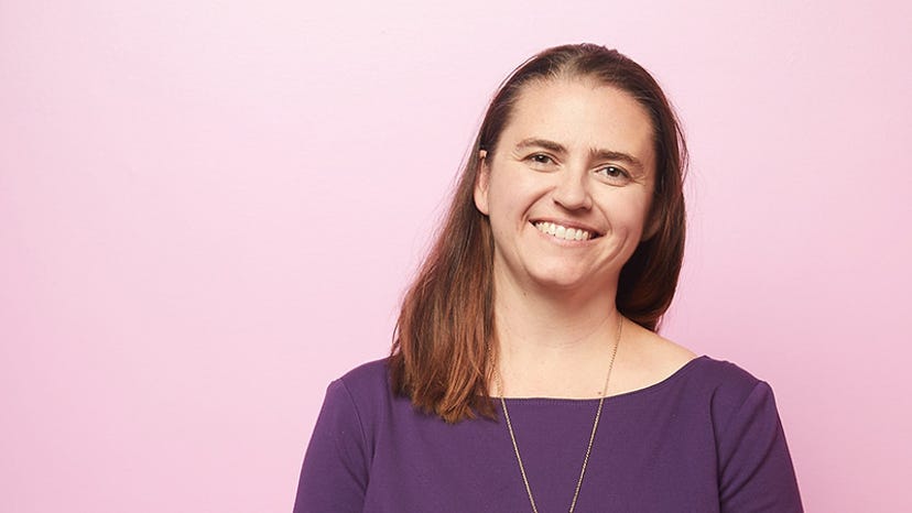 A headshot of Lyndsay Pearson. She is wearing a purple top against a pink background.