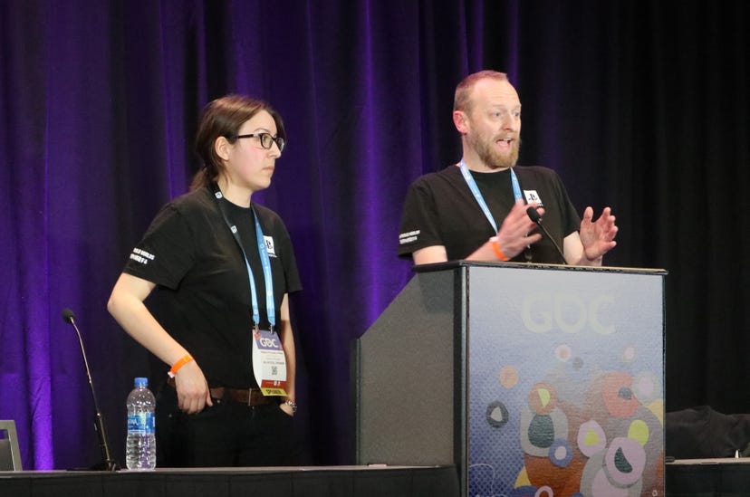 two adults stand on a stage behind a podium; the person on the right waves his hands while speaking into a microphone