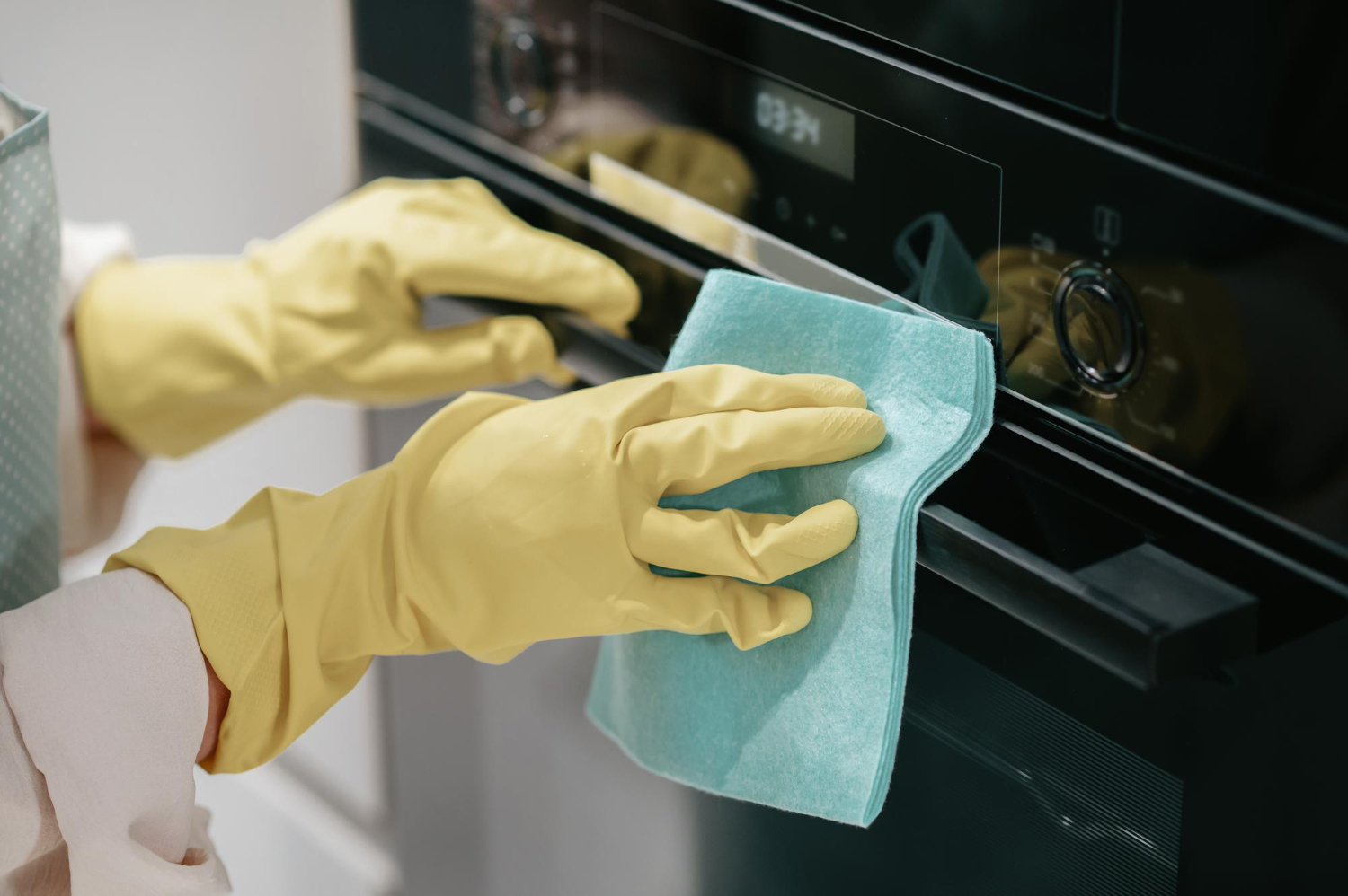 Mãos com luvas amarelas limpando painel de forno de fogão preto com pano azul. 