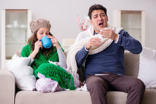 Two people sitting on a couch wrapped in blankets while sneezing and drinking a hot beverage