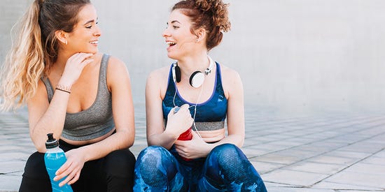 Two young girls, sitting back to back in exercise clothes, smiling and holding water bottles