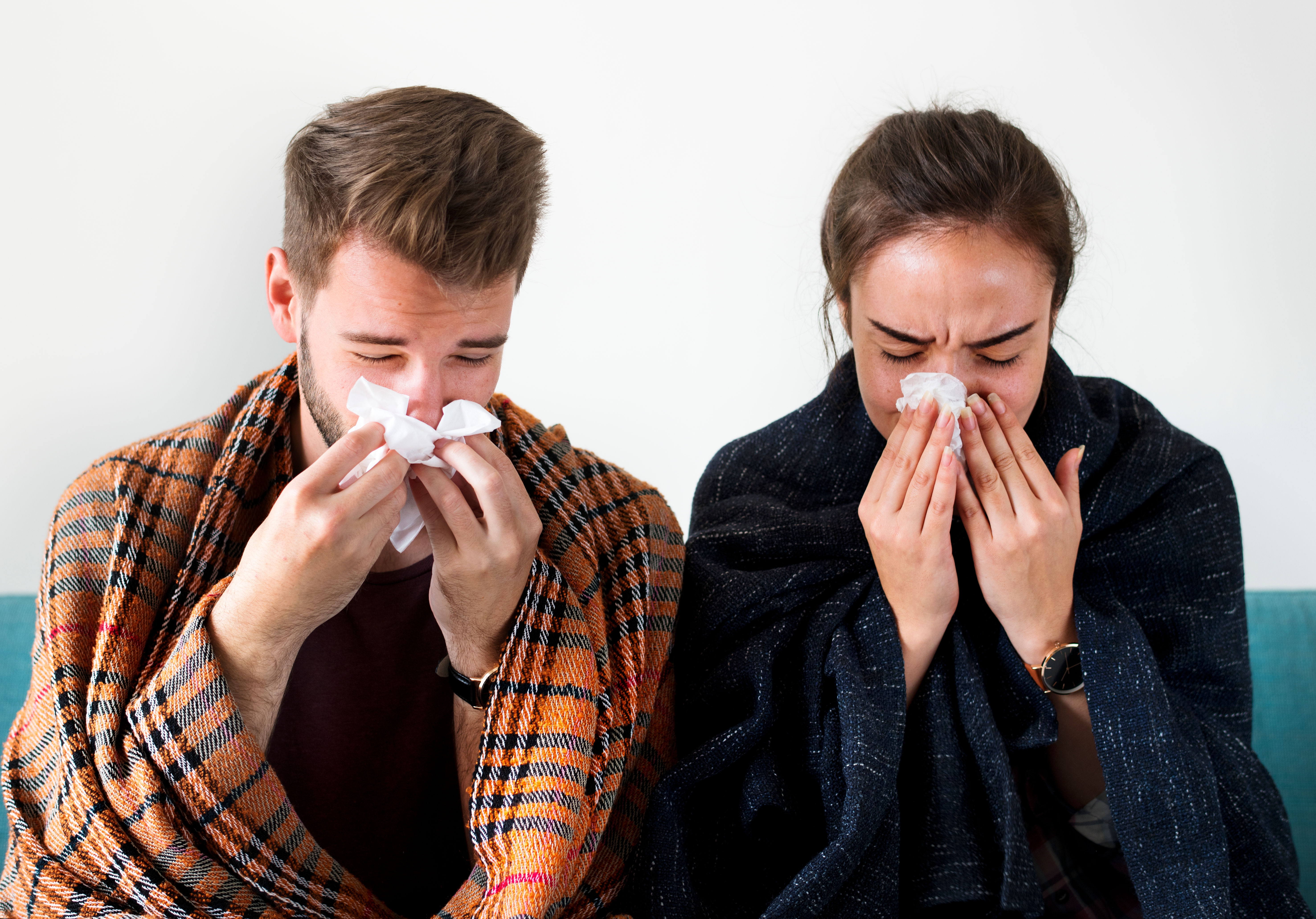 A couple with cold and flu blowing their noses into tissues.