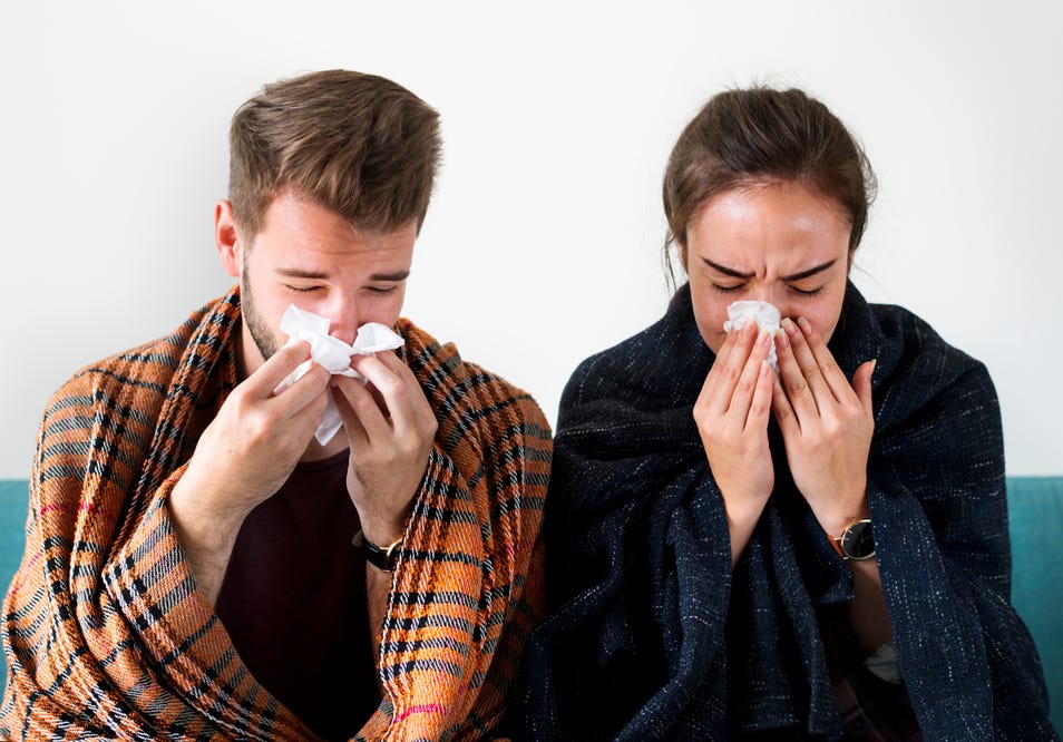 A couple with cold and flu blowing their noses into tissues.