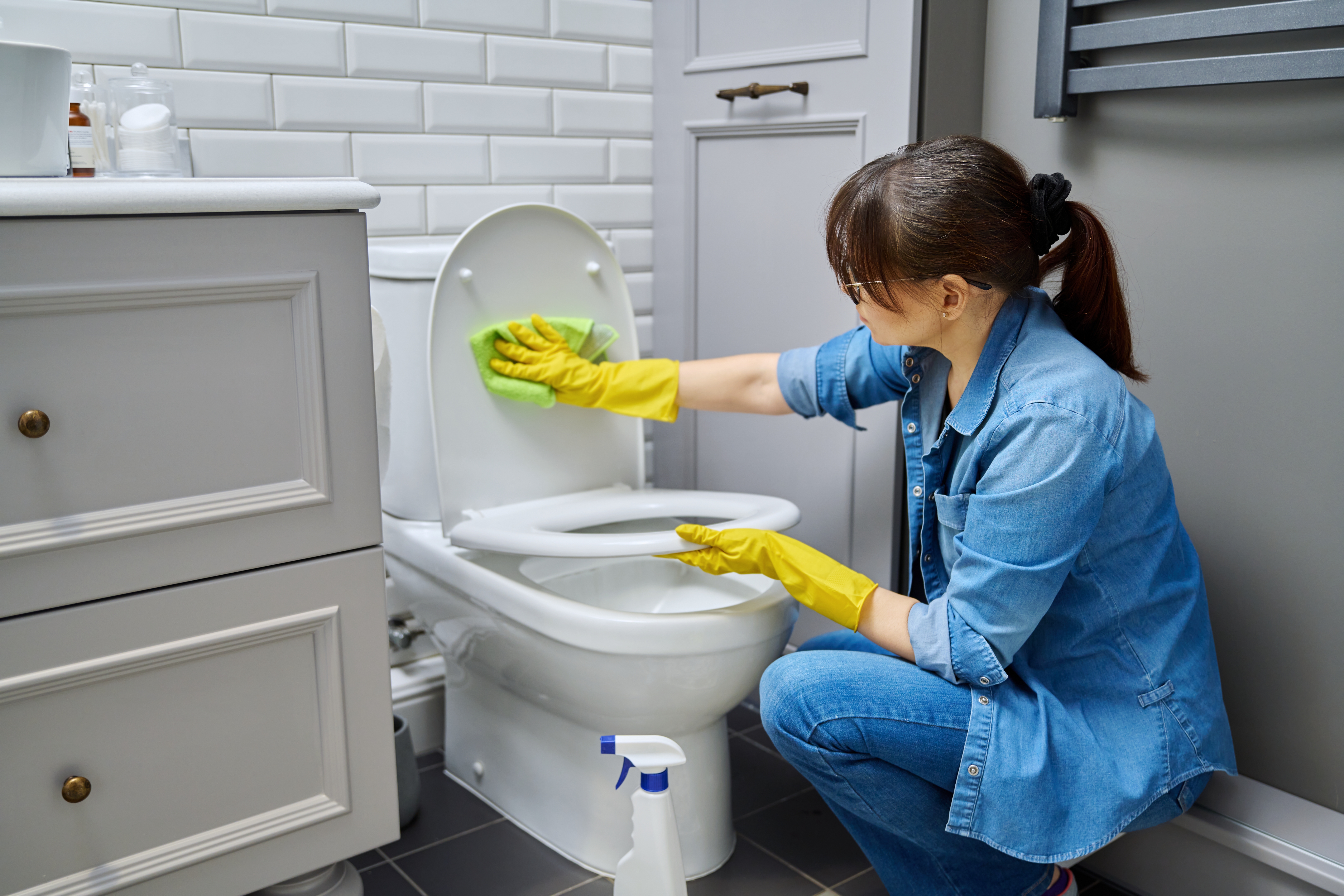 A woman wearing yellow rubber gloves, cleaning the toilet seat with a cloth.