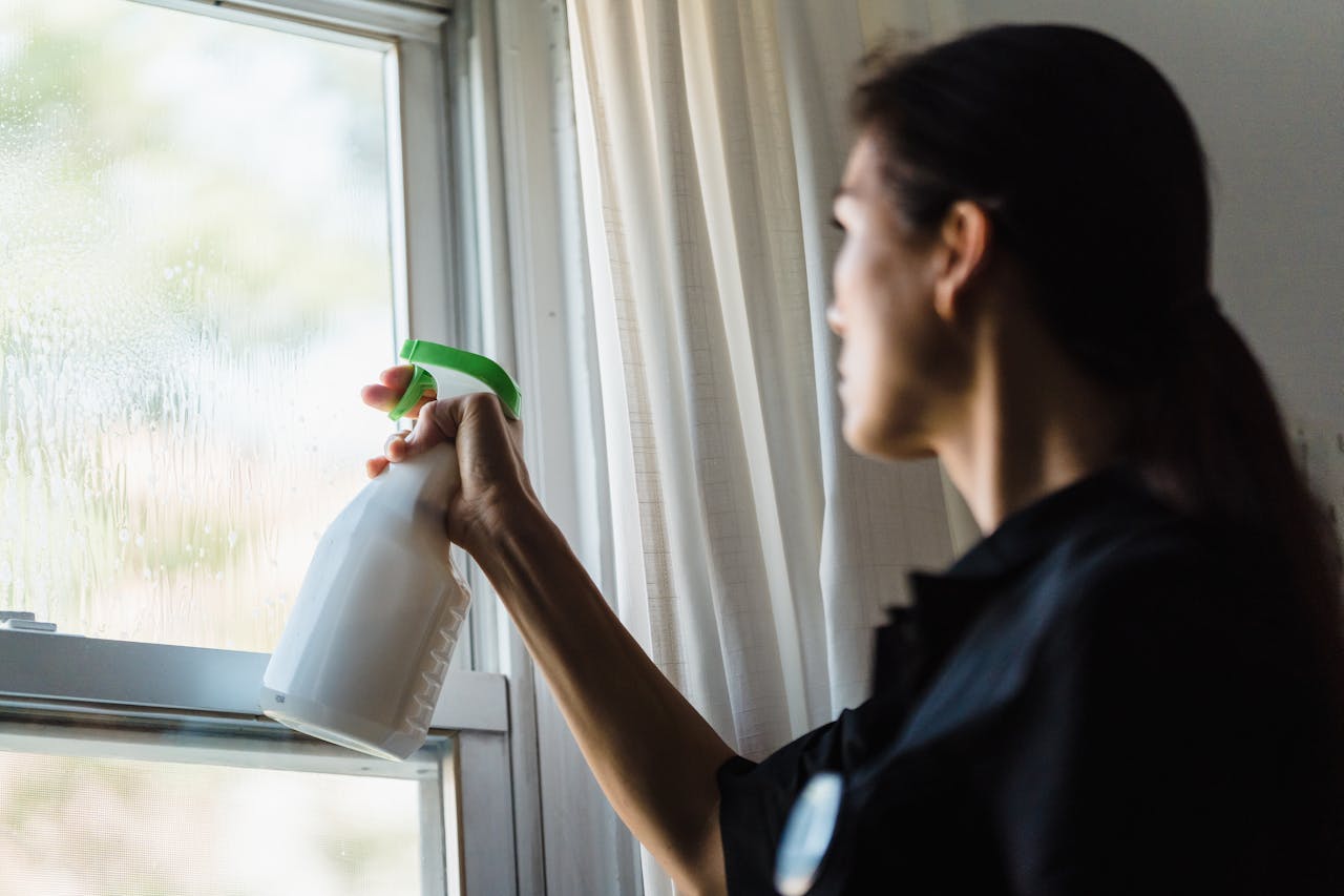 Mulher borrifando produto de limpeza em vidro, com a mão segurando o spray em direção à janela