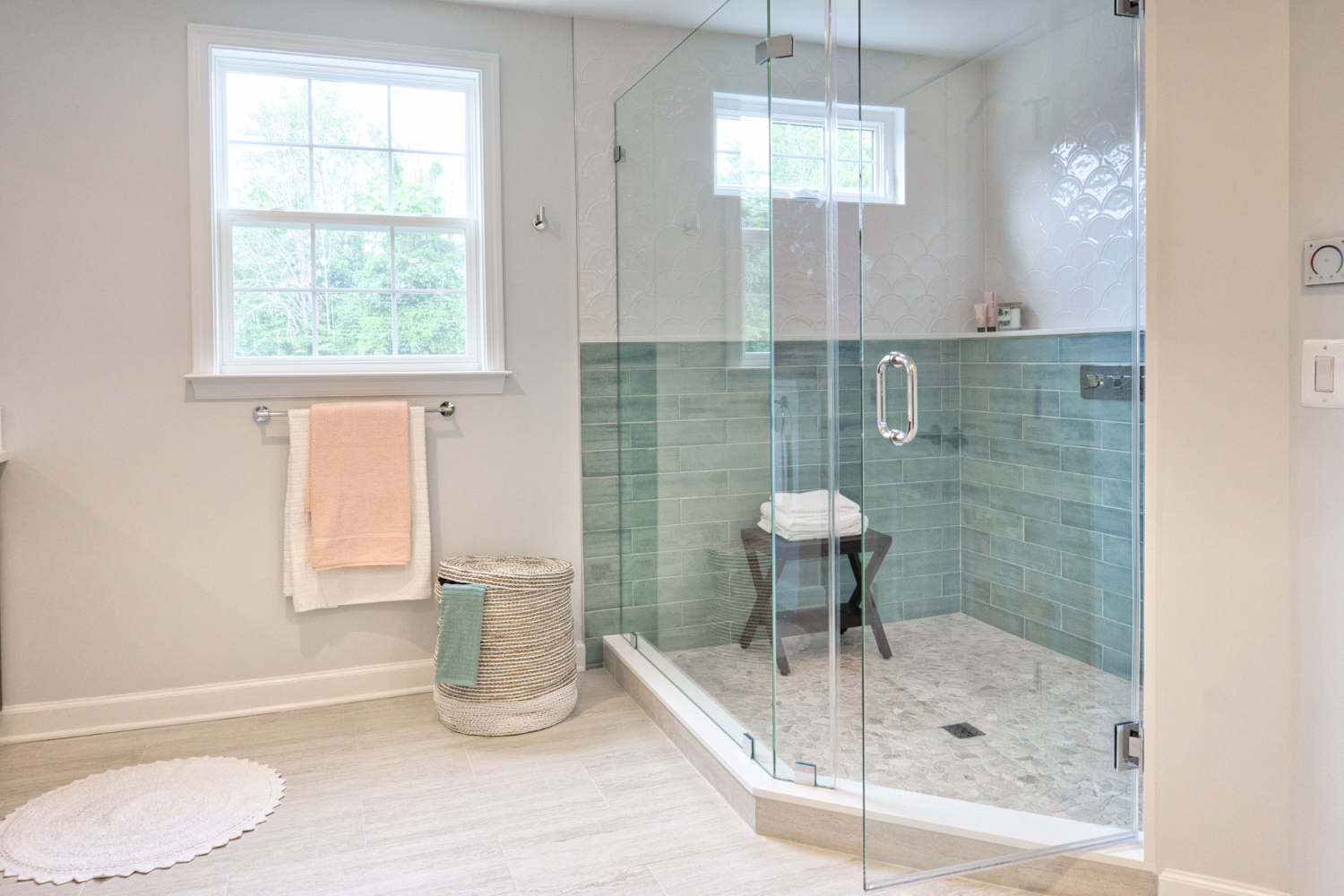 Banheiro moderno com chuveiro de vidro transparente, paredes com azulejos verdes e brancos, toalhas penduradas, cesto de roupa e banco interno no box.