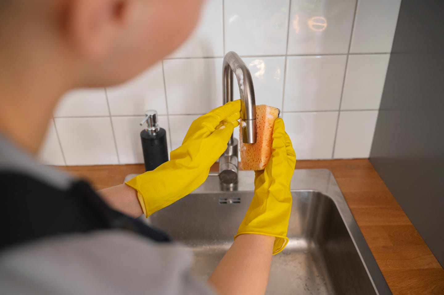 A person wearing yellow gloves is cleaning the kitchen faucet with a sponge