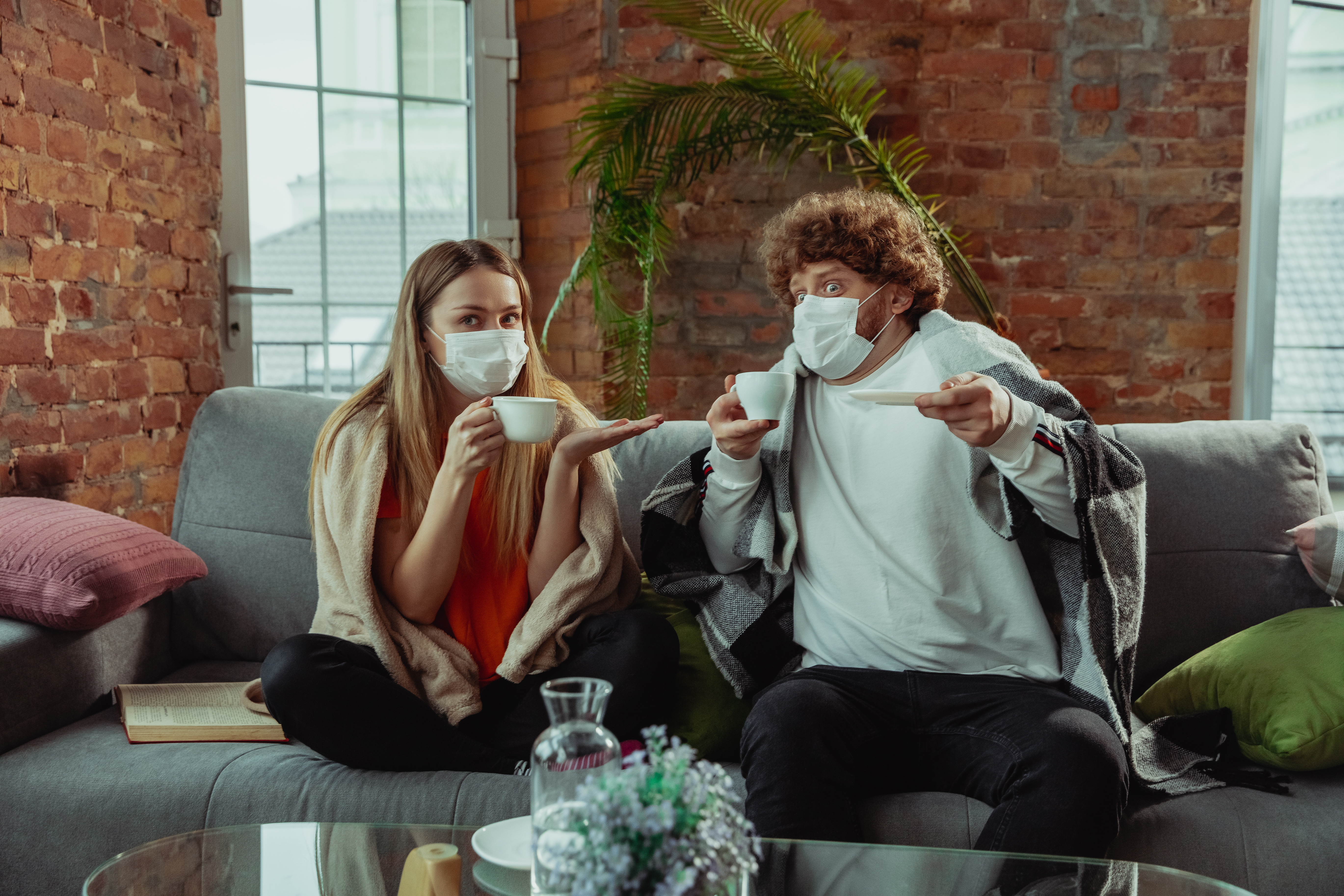 Two people wearing face masks and blankets sit on a couch with cups. 