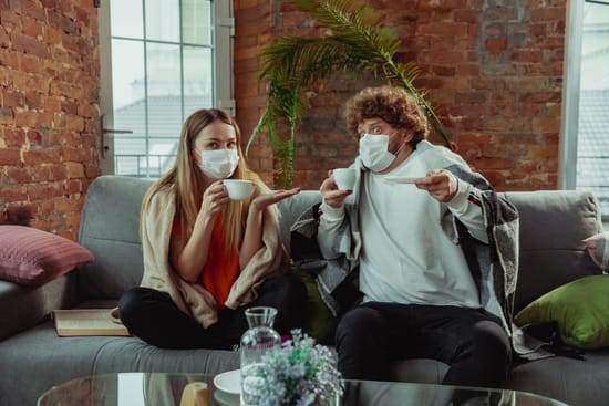 Two people wearing face masks and blankets sit on a couch with cups.