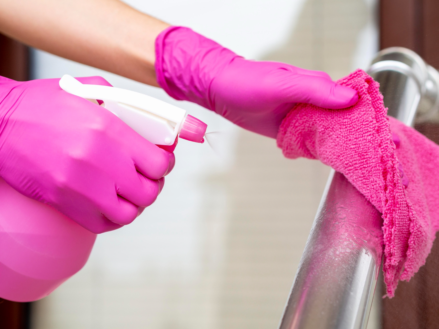 Pessoa com luvas limpando superfície metálica com pano rosa, borrifando produto de limpeza.