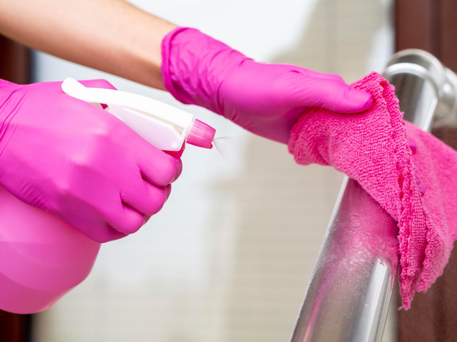 Pessoa com luvas limpando superfície metálica com pano rosa, borrifando produto de limpeza.