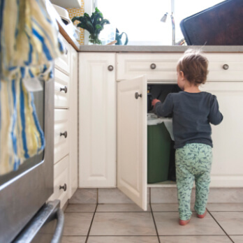Young child in a kitchen with an open cupboard tossing trash into the garbage