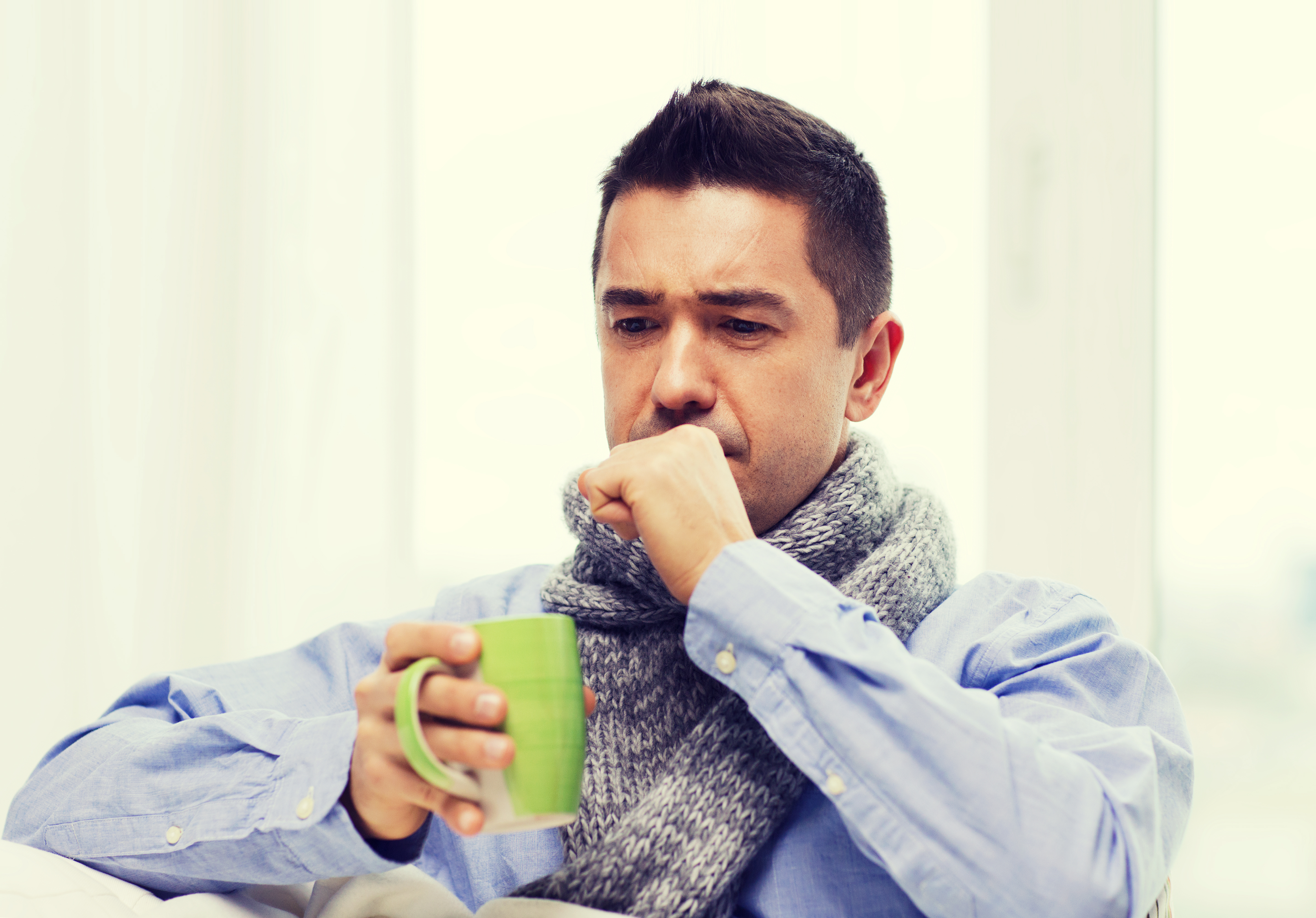 Man in a knitted cloth coughs into his fist while holding a warm drink. 